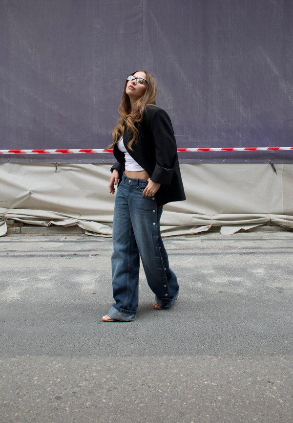 Femme portant des lunettes de soleil, un blazer noir, un crop top blanc et un jean à jambes larges, marchant dans une rue avec une barrière de chantier derrière elle.