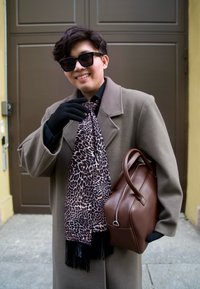 Borsa a mano in pelle marrone, cappotto beige, guanti neri e sciarpa con stampa leopardata e frange. La modella indossa occhiali da sole e sorride.
