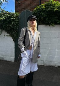Šedý pruhovaný oversize blazer přes bílé tričko, v kombinaci s bílými šortkami a vysokými černými botami. Doplněno černou čepicí a st�říbrnými šperky.