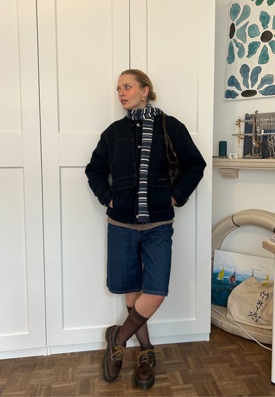 Chaqueta de mezclilla oscura sobre un suéter claro, pantalones cortos de mezclilla azules, bufanda a rayas, zapatos marrones y un bolso marrón. El fondo presenta armarios blancos.