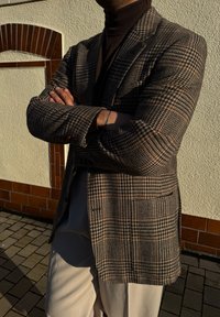 Blazer com padrão de houndstooth marrom e corte tailored, sobreposto a um suéter de gola alta escuro, combinado com calças de tom claro.