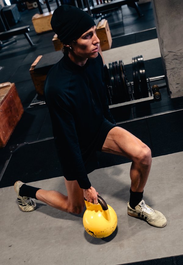 Mladi moški v črnih športnih oblačilih drži rumeno utež kettlebell, medtem ko kleči v telovadnici, kjer so v bližini uteži in lesene škatle.