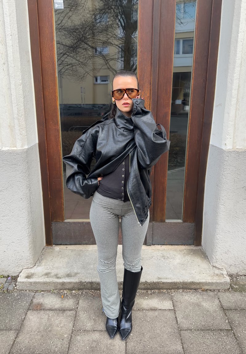 Femme portant une veste en cuir noire surdimensionnée, un pantalon rayé, des bottes noires et de grandes lunettes de soleil, debout sur le trottoir à côté d'une porte en bois.