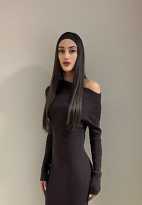 Femme avec de longs cheveux noirs et raides, portant un serre-tête, vêtue d'une robe noire ajustée et décolletée, debout devant un mur beige uni.