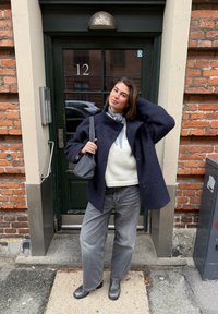 Temno modri volneni plašč čez svetel pulover, sive široke jeans hlače in črni gležnjarji. Drži majhno sivo torbico pred opečno zgradbo.