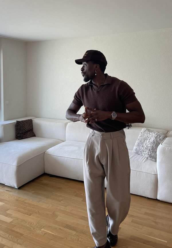 Uomo con camicia marrone scuro, pantaloni beige e cappello, in piedi in una stanza con pavimento in legno chiaro e divano angolare bianco con cuscini.