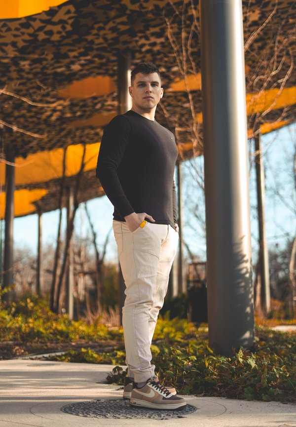 Maglietta nera a maniche lunghe, pantaloni slim-fit beige e sneakers grigie con dettagli marrone chiaro. Sullo sfondo, un tendaggio a motivi e vegetazione.