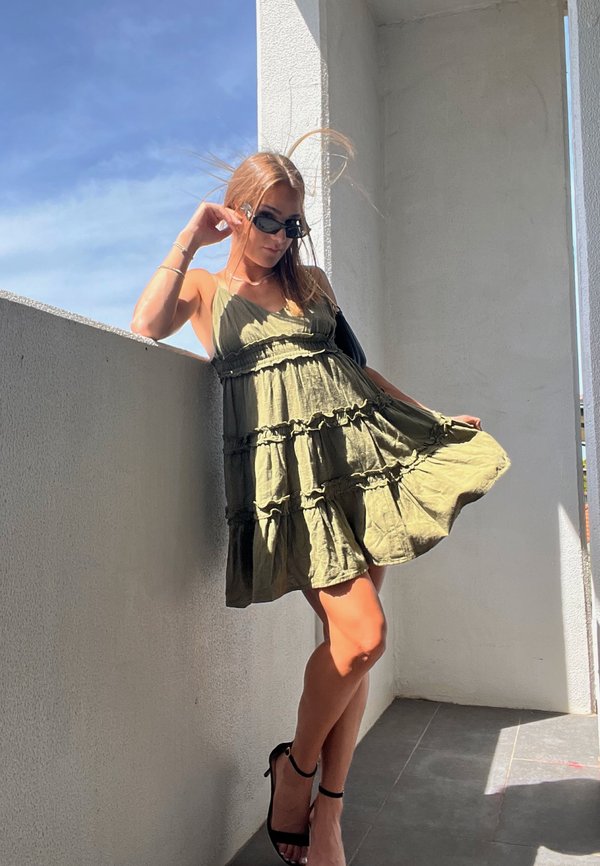 Joven mujer con gafas de sol posa en el balcón vistiendo un vestido sin mangas de color verde oliva y sandalias negras de tacón alto bajo la luz del sol.