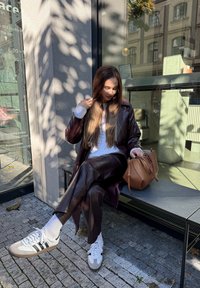 Ženska v rjavem usnjenem plašču in hlačah sedi na zunanji klopi in ureja lase, ob njej na klopi je svetlo rjava torbica.