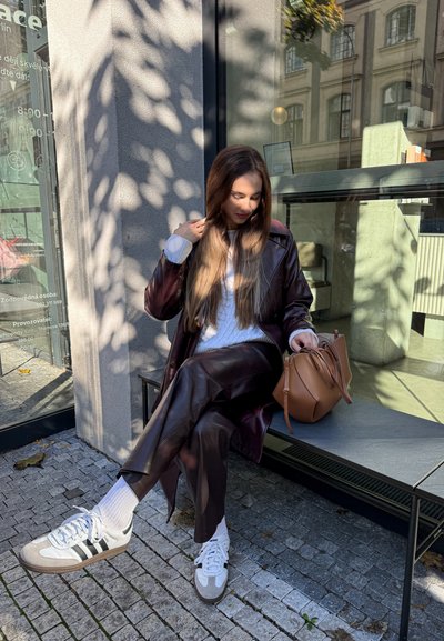 Mujer con abrigo y pantalones de cuero marrón sentada en un banco al aire libre ajustándose el cabello, con un bolso beige en el banco a su lado.