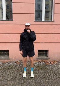 Zwarte sportjas met capuchon en ritssluiting, gecombineerd met zwarte shorts, blauwe sokken en lichte sneakers, tegen een roze muur.