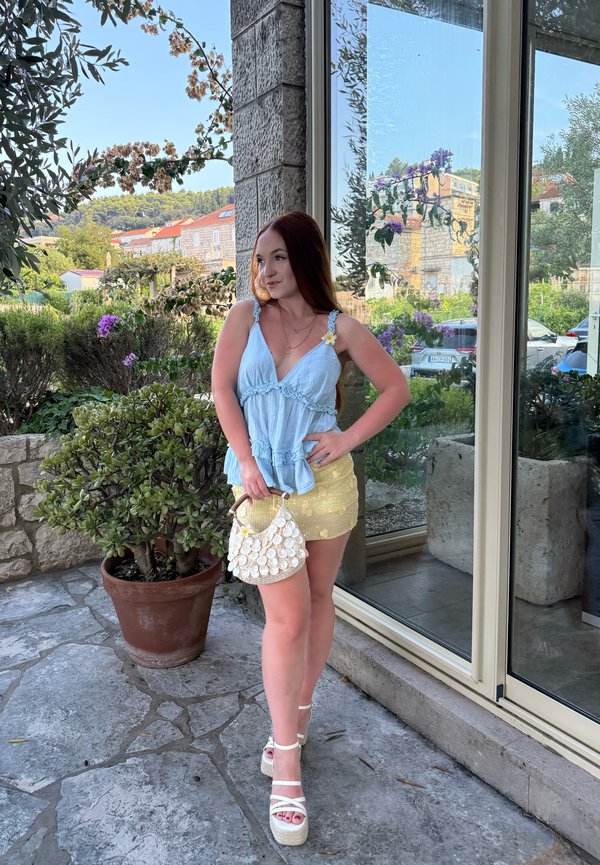 Jeune femme en haut bleu et jupe jaune tenant un sac à main en forme de coquillage, debout sur une terrasse en pierre à côté d'une fenêtre en verre avec des plantes et des bâtiments à l'extérieur.