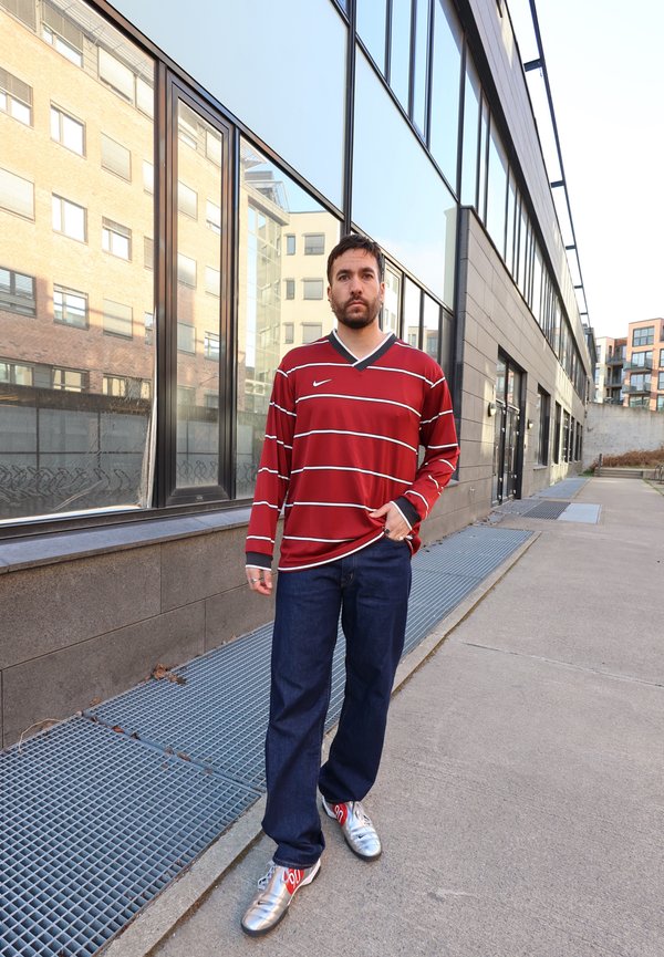 Man met kort donker haar en baard, gekleed in een rood gestreept shirt met lange mouwen, donkere jeans en zilveren sneakers, staand op een stadsstoep.