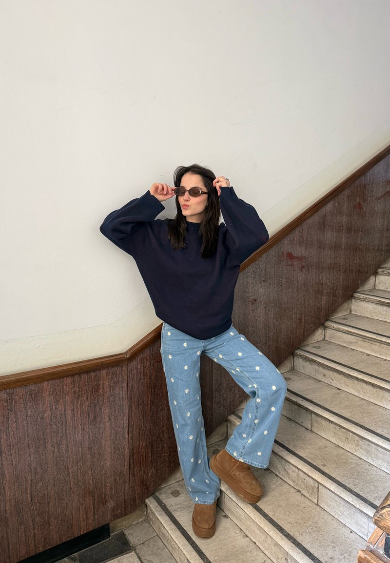 Kvinde justerer små solbriller, iført mørk trøje, blå jeans med hvidt blomstermønster og brune plateau-sko, stående på en trappe.