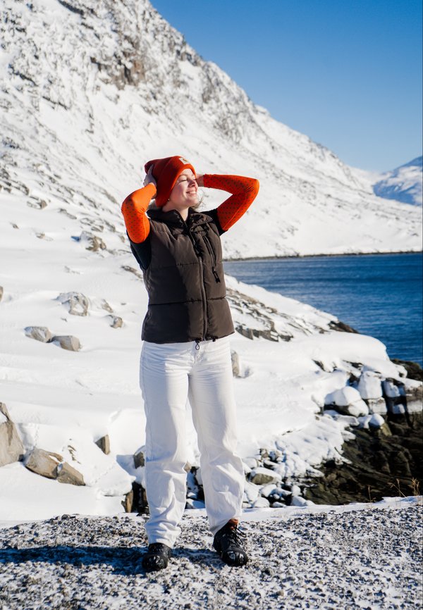 Gilet nero imbottito, maniche lunghe arancioni con motivi, pantaloni bianchi, berretto rosso, in piedi su una riva rocciosa con neve e acqua sullo sfondo.