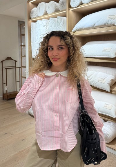Mujer con cabello rizado que lleva una camisa rosa de rayas y sostiene un bolso negro, de pie en un pasillo de una tienda con almohadas blancas exhibidas en estantes.
