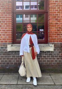 Een vrouw staat naast een bakstenen muur, gekleed in een witte blouse, beige rok en een bruine riem, terwijl ze een crèmekleurige tas vasthoudt, met een rode sjaal en witte sneakers.