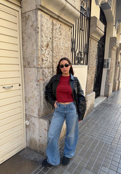 Mujer con gafas de sol, top rojo corto, chaqueta de cuero negra y jeans de pierna ancha, de pie contra una pared de piedra en la acera urbana.