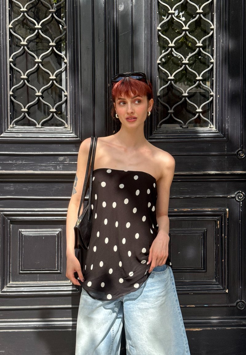 Jeune femme aux cheveux rouges courts portant un top noir sans bretelles à pois, un jean bleu clair, des lunettes de soleil sur la tête, debout devant des portes décoratives foncées.