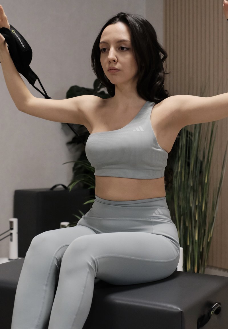 Jeune femme en vêtements de sport gris clair assise sur un appareil d'exercice, bras levés, tenant un masque facial noir, à l'intérieur avec des plantes à proximité.