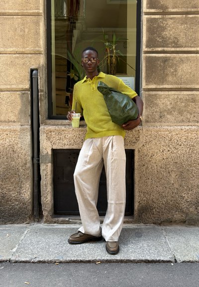 Bolso verde de gran tamaño, polo amarillo de punto, pantalones beige de pierna ancha, zapatos marrones; sosteniendo una bebida, posando al lado de una pared de piedra y una ventana.
