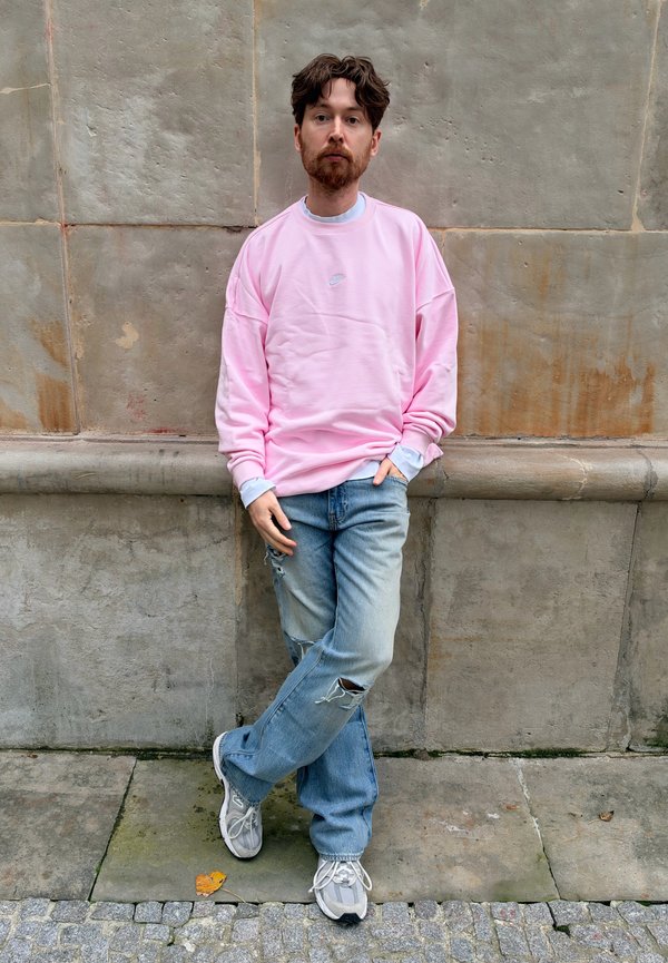 Hombre con cabello castaño y barba, vestido con una sudadera rosa, vaqueros azules rotos y zapatillas deportivas, apoyado contra una pared de piedra con las piernas cruzadas.