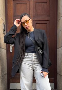 Giacca nera oversize su un top blu navy, jeans a gamba larga grigio chiaro e occhiali con lenti gialle. Accessoriata con una collana di perle e una borsa a tracolla.