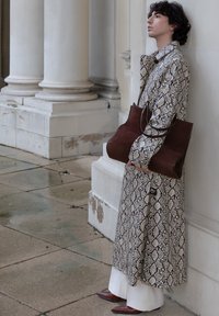 Personne portant un long manteau à imprimé serpent, tenant un grand sac cabas en cuir marron, debout devant un bâtiment blanc à colonnes.