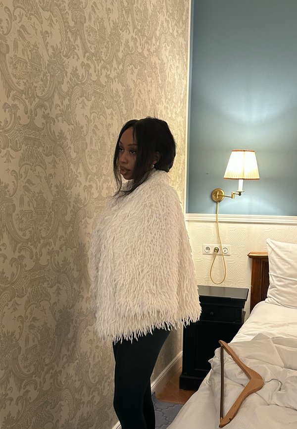 Vrouw met een witte, pluizige sjaal staand naast een beige muur met een patroon, naast een bed met een houten hanger erop, in een zacht verlichte kamer.