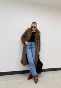 Καφέ ψεύτικο παλτό, μαύρη μπλούζα, ανοιχτό μπλε τζιν, κόκκινες γόβες με μυτερή μύτη και μαύρα γυαλιά ηλίου; μακρύ παλτό με υφή και λεπτομέρειες από φούντες.