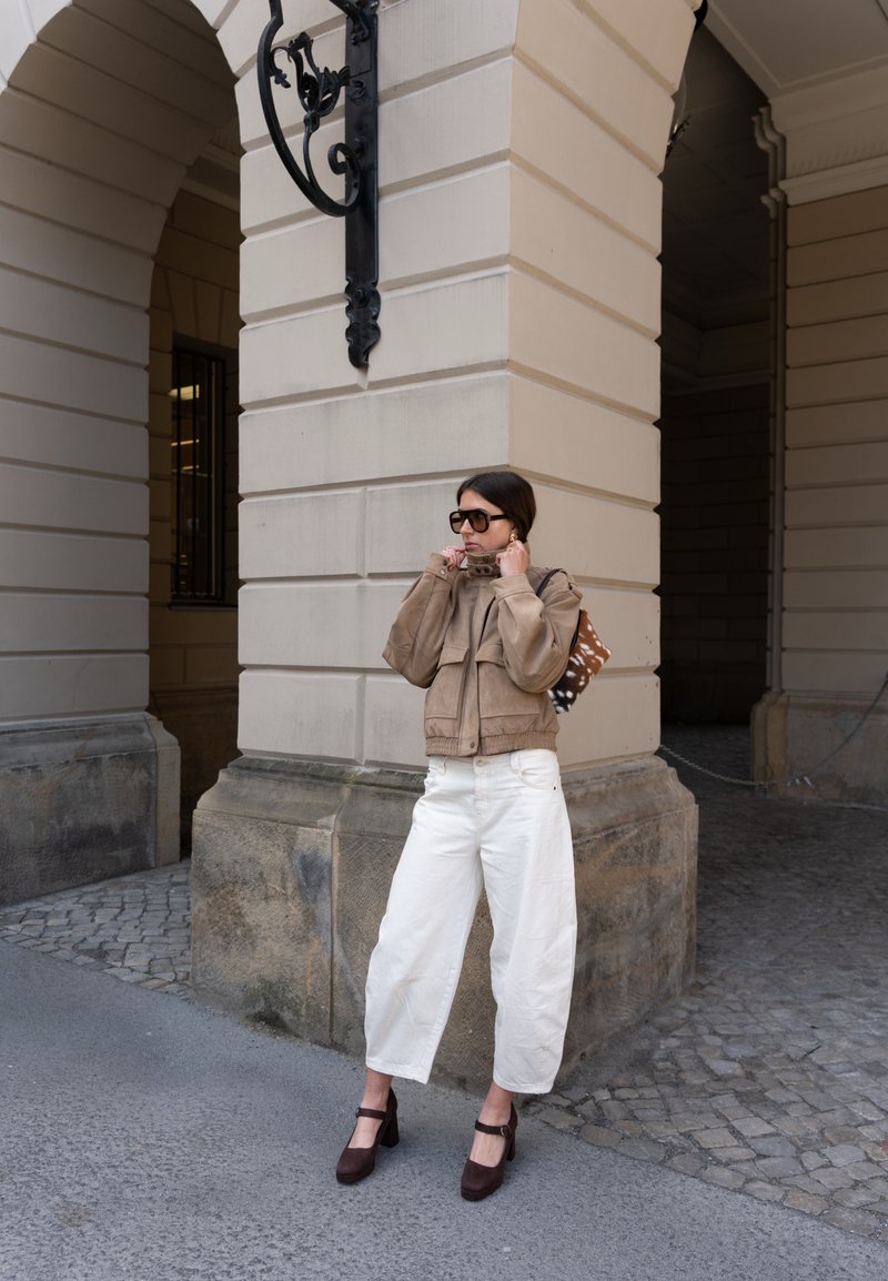 Femme vêtue d'une veste beige, d'un pantalon blanc à jambes larges, de talons marron et de lunettes de soleil, debout près d'une arche en pierre en ajustant le col de sa veste.