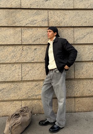 Chaqueta bomber negra, suéter texturizado crema, jeans holgados grises y zapatos negros brillantes. Bolsa de lona marrón en el suelo contra una pared de piedra.