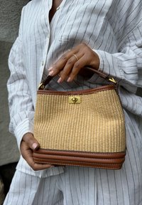 Borsa di paglia intrecciata con base e tracolla in pelle marrone. Presenta una chiusura in oro e una forma strutturata, evidenziando una superficie testurizzata.