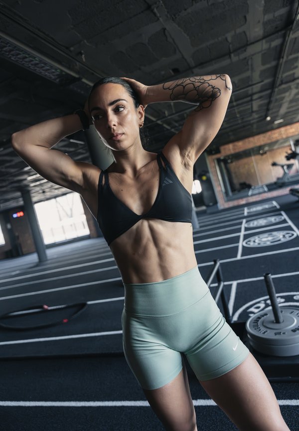Sportliche Frau mit Tätowierung streckt die Arme hinter dem Kopf in einem Fitnessstudio mit Gewichtsscheiben und Trainingsgeräten im Hintergrund.