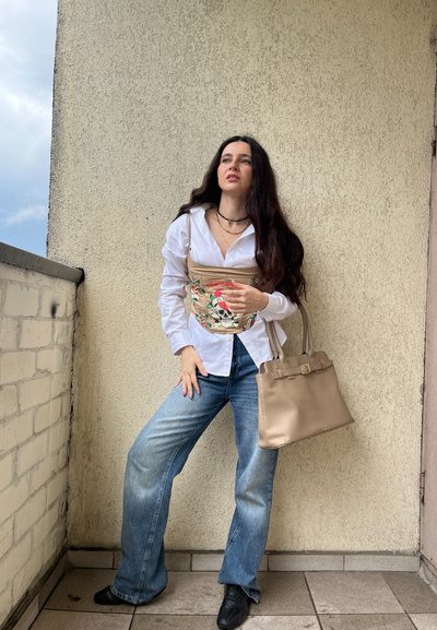 Bolso beis con diseño estructurado y detalle de hebilla. El modelo lleva una camisa blanca de botones y jeans de pierna ancha en azul claro, de pie junto a una pared.