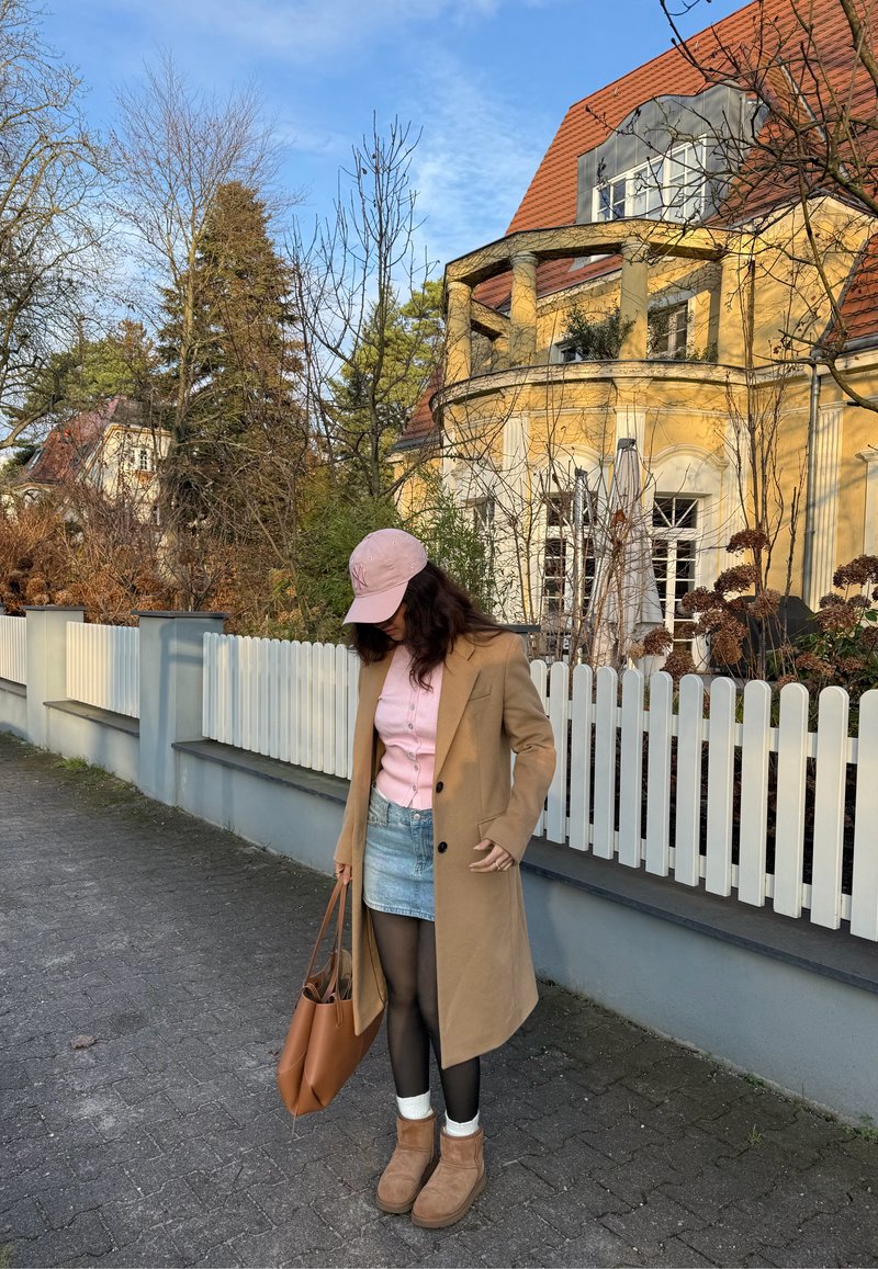Long manteau beige, chemise rose à boutons, jupe en jean, bottes marron et casquette rose. Sac fourre-tout marron, collants noirs, avec un fond de clôture blanche en bois.