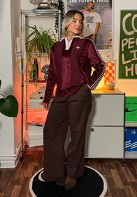 Mørkerød sateng, langermet topp med hvit krage og sidestriper; kombinert med brune vide bukser. Tregulv og moderne dekor i bakgrunnen.