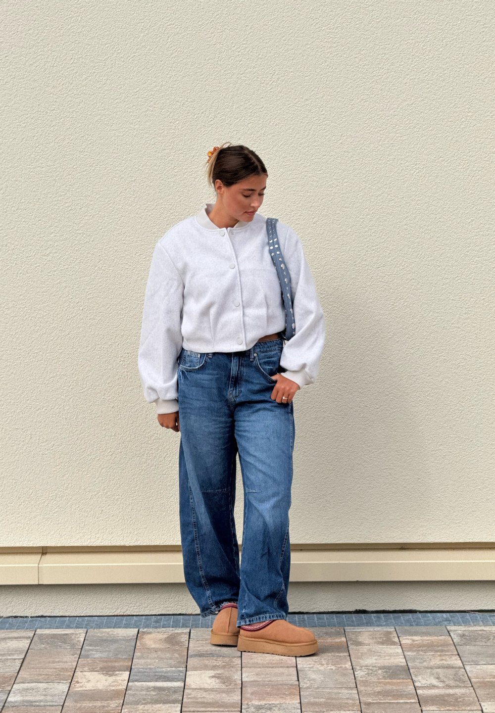 Giacca bianca, soffice e corta; jeans blu a vita alta; scarpe slip-on color sabbia; tracolla grigia con borchie; sfondo neutro.