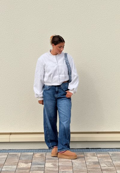 Chaqueta corta blanca y esponjosa; jeans azules de cintura alta; zapatos de lona color beige; bandolera gris con tachuelas; fondo neutro.