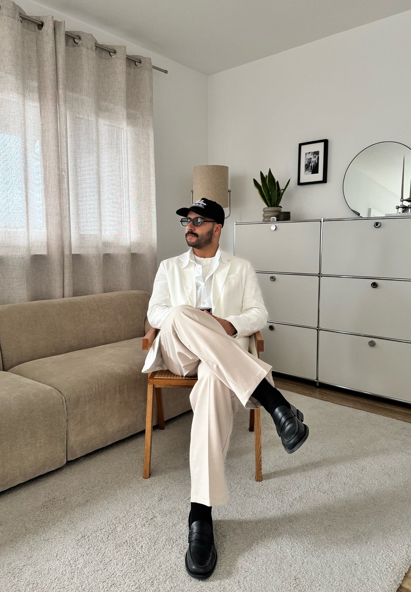 Man in white blazer, light pants, black cap, and glasses sitting with legs crossed on a wooden chair in a beige living room.