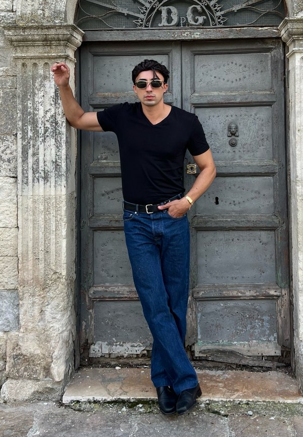 Hombre con camisa negra, vaqueros azules y gafas de sol apoyado en una puerta de madera vieja con marco de piedra, una mano en el bolsillo, un brazo levantado.