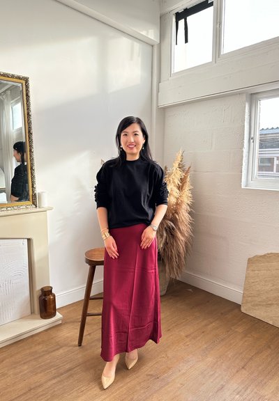 Sudadera negra, falda de satén burdeos, tacones puntiagudos nude. Accesorios que incluyen un reloj y pulseras. Texturas suaves, diseño minimalista.