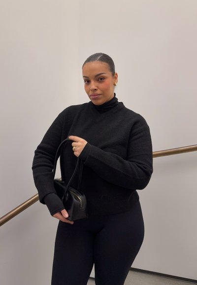 Mujer con el cabello recogido, vistiendo un suéter negro y leggings, sostiene un pequeño bolso negro, de pie junto a una pared beige y una barandilla.