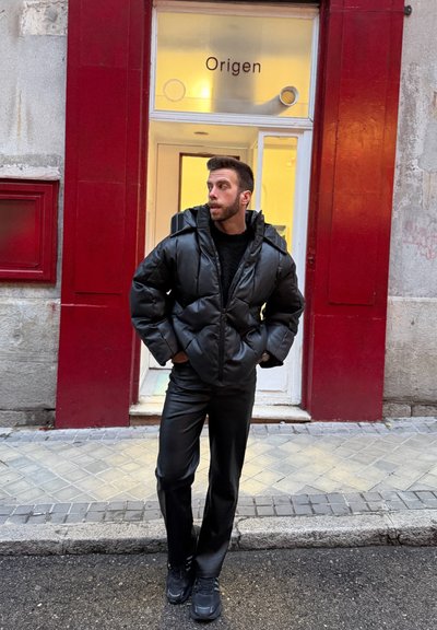 Hombre con chaqueta de plumón negra y pantalones de pie en una acera, mirando de lado frente a un edificio rojo y gris con un cartel que dice "Origen" sobre la puerta.