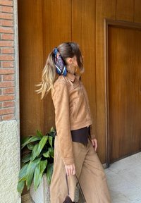 Brązowa zamszowa kurtka z ozdobami w postaci ćwieków, krótki fason, zestawiona z pasującymi szerokimi spodniami. Włosy związane niebieską apaszką z wzorem.