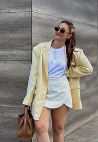 Žuti oversized sako, bijela majica i svijetla zavojna suknja. Smeđa torba s zlatnim detaljima. Kratka, ravna teksturirana kosa.