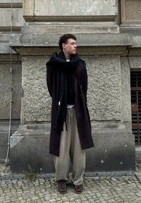 Cappotto nero lungo, pantaloni beige oversize, sciarpa nera, scarpe scure. Parete grigia testurizzata e superficie di ciottoli sullo sfondo.