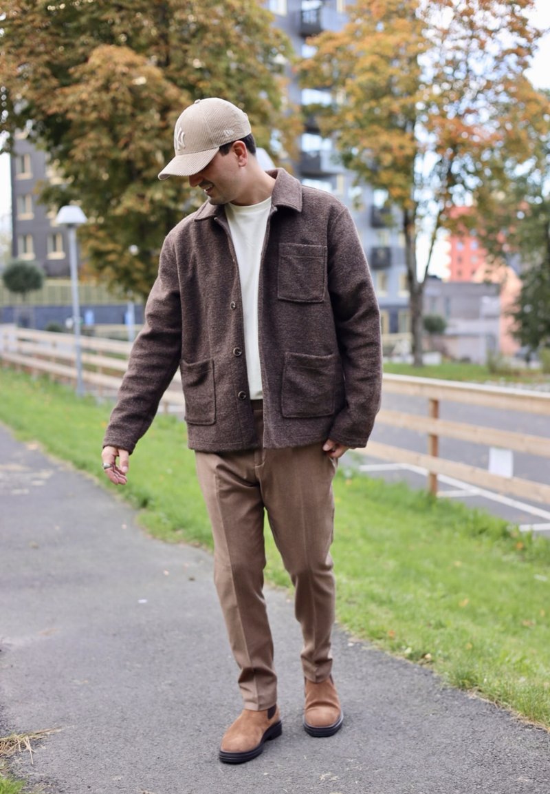 Hnědá vlnená bunda s límečkem a dvěma předními kapsami, kombinovaná s béžovými kalhotami a hnědými kotníkovými botami, umístěná v přírodním prostředí.