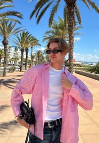 Lichtroze gestreepte oversized shirt, witte crew neck t-shirt, zwarte jeans en een zwarte handtas tegen een achtergrond van palmboom en blauwe lucht.