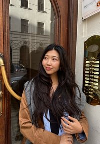 Młoda kobieta z długimi ciemnymi włosami, ubrana w nałożone na siebie kurtki, stoi obok drewnianych drzwi z mosiężną klamką i panelem domofonu do mieszkania.